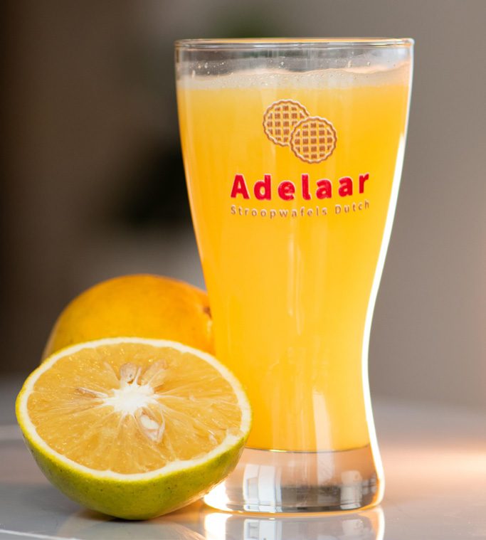 Vaso de jugo de naranja Adelaar recién exprimido junto a naranjas partidas